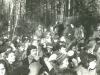 2nd Jewish Song Festival, Succot, 1979. More participants attended this time. There are a lot of young people in the audience. The symbolic sukkah served as a hall for the festival. Pine tree branches hang on the ropes. Policemen are standing behind the branches (they can be seen in the background on the left side). Refuseniks are scattered throughout the audience: at the wall of the sukkah (in the center)  Sasha Kremen (boy), next to him are his mother, Galya Kremen, and Galya Gurevich. In the second row (to the left), sitting and attentively looking at the stage is Kira Razgon a singer and attendee of all the festivals. In the first row (to the right), in profile (without a hat) is Lena Gurevich. Moscow, Ovrazhki forest, Succot, 1979. co RS